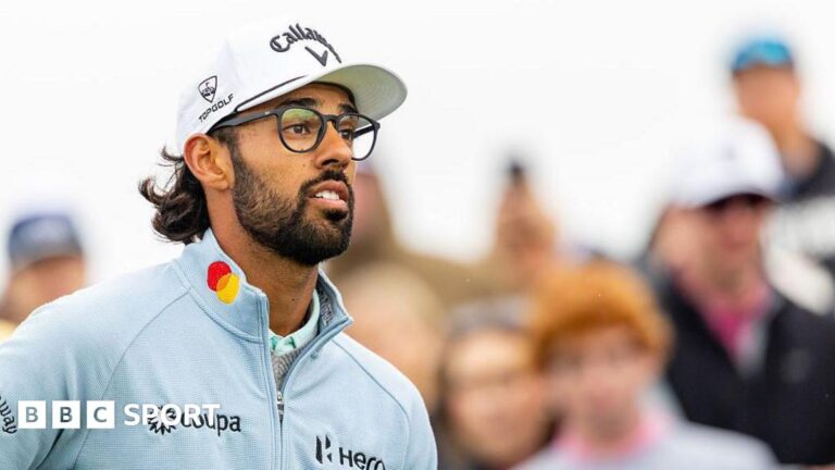 Akshay Bhatia looks on at the ninth tee. He has a black beard and is wearing black-rimmed glasses, a white cap and a light blue zip-up top. People in the crowd can be seen behind him.