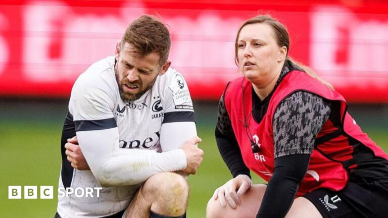 Elliot Daly (left) kneels on the pitch with his arms bent across his knee clutching his left elbow, with a grimace on his face during Saracens' defeat by Bath on 4 April