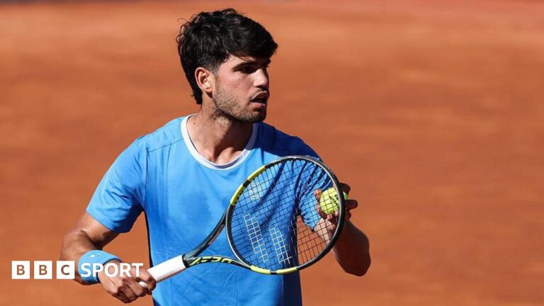 Carlos Alcaraz is wearing a blue t-shirt and holding a tennis ball to his racquet