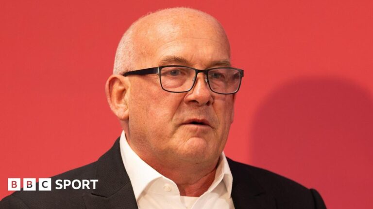 A close-up of Richard Collier-Keywood in dark jacket and open white shirt in front of a red background