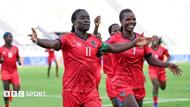 Malawi striker Tabitha Chawinga runs towards the touchline with arms spread wide to either side of her body to celebrate a goal at the 2026 Women's Africa Cup of Nations in Morocco. She wears all-red kit with green and black trim and is surrounded by a number of smiling team-mates who are running in her wake across the lush green pitch