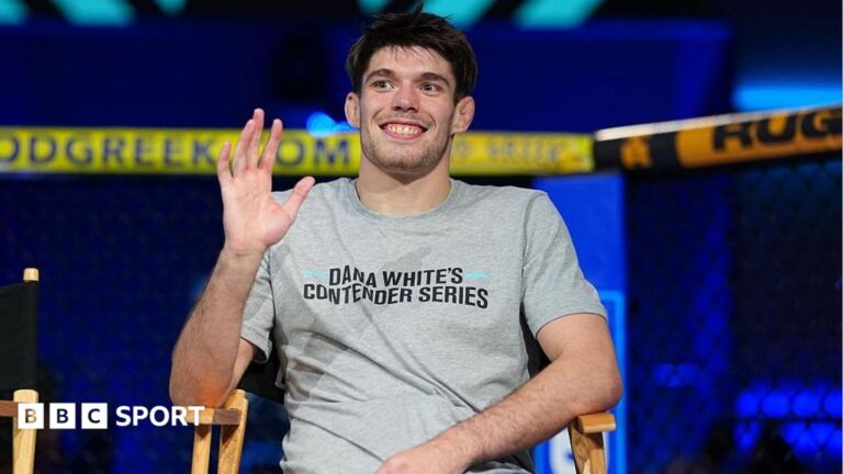 George Staines is smiling and waving as he wears a grey t-shirt